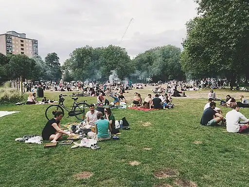 Diverse crowd of people sit and picnic in an urban park, some have brought barbecue grills