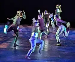 Group of dancers in black and metallic colors in exaggerated running poses pointing in various directions