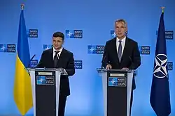 Two men in business suits stand at silver podiums, with a blue and yellow Ukrainian flag to their left and a blue and white NATO flags to their right.