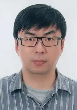 Man in gray pinstriped shirt standing with right hand raised in front of a podium and microphone stand