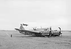 A twin-engined World War II–era monoplane lying on its belly on grass. Both of its propellers are twisted and another aircraft is flying near the ground behind it.