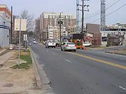Ground-level view of a mid-sized street in an urban area, with one lane closed in the direction headed away from the viewer. In the far background are high-rise office and residential buildings, while the near background features a construction site with a large crane against an overcast sky on a winter day.