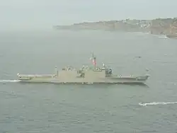 A large grey warship underway. Flags are being flown from the ship's mast, and white-uniformed people line the decks. Brown cliffs line the horizon for the right two-thirds of the image