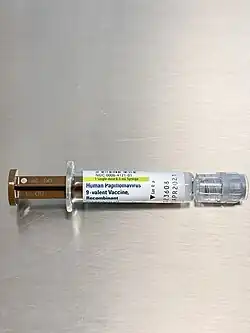 A photo of a short syringe with a brown plunger and a luer-lock cap resting on a brushed metal surface. The a label is on the glass syringe, it reads Human Papilloma Virus, 9-Valent Vaccine, Recombinant. Other text informs the reader of the lot number and expiry date.