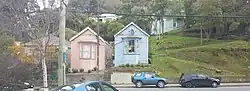 Two almost identical small wooden houses next to each. A pink one and a blue grey one. Set up from the street with a concrete retaining wall. A steep grassy bank is to the right. Parked cars and street posts in the foreground.