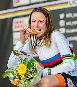 Annemiek van Vleuten wearing a jersey with rainbow stripes, biting a gold medal and holding flowers.