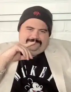 A straight-on bust photograph of a 34.6-year-old white man with a dark mustache; he is wearing a black beanie, Mickey-Mouse t-shirt, and beige blazer while looking into the camera with a contemplative pose