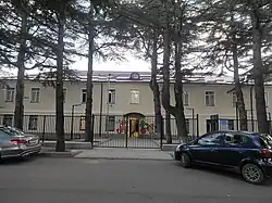 A two-story, pale yellow building with a dark roof, seen from across a street through a line of tall, slender trees. A green metal fence separates the building from the sidewalk.