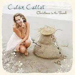 Colbie Caillat sitting on the sand, with a starfish in her hand, next to a sandman, in the beach.