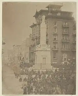 An antique photograph depicting a city square with a stone monument and a large number of soldiers at rest