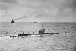 A submarine sailing on the surface of the ocean, while in the background is a large warship and a smaller vessel.