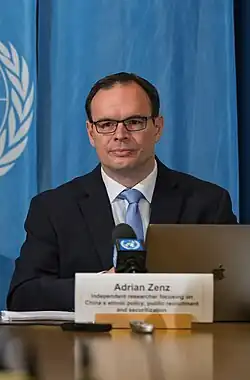 Adrian Zens wearing a white shirt, light blue tie, and dark jacket, sitting behind a table, laptop, microphone, remote controls, and name tag in front of him, and United Nations logo behind him on blue curtain