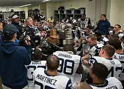 A man speaks to American football players in uniform in a locker room in front of a large trophy.