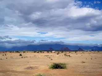 Daytime view of an almost lifeless expanse, dry rocks and sand marked only by the odd lone shrub. The dry terrain reaches to a chain of mountains in the far distance, near the horizon. A bank of clouds soars above the void, but it does not appear to hold the promise of rain. A far darker, larger, more turgid cloud bank sits above the and distant mountains, above the horizon.