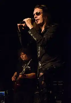 Aleš Brichta standing onstage in black clothes and wearing dark sunglasses, singing into a microphone