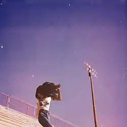 A young woman standing on football bleachers in front of a blue sky and stadium light