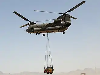 Colour photo of a green twin-rotored helicopter in flight with a yellow front loader suspended below the aircraft by a rope