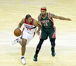 At left man carrying the ball wearing a white Wizards uniform, man at right guarding him wearing a red headband and dark green Bucks uniform