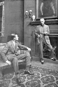 young man seated talking to older man standing by large fireplace