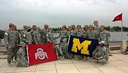 Two groups of soldiers in camouflage uniforms staring at each other on a waterfront; the left group carries a red flag with a silver "O" and the words "OHIO STATE" on it; the right group carries a blue flag with a yellow "M" on it.
