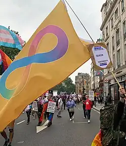 Autisitic pride flag waved in a street with other protesters, the flag has a mask witch has a paper witch has text "autistic, queer, solidarity" above and below the text is a bi flag drawn