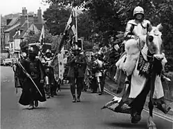 An armoured and mounted man leads a small party, similarly dressed in mediaeval attire, along a road.