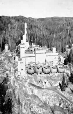 Boxy buildings and the mine's head shaft behind ten large tanks, surrounded by trees