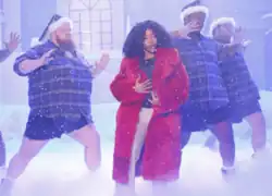 A woman wearing a red fur coat, who is surrounded by stout male dancers wearing shorts, checkered shirts, and Santa hats. They are on a winter-themed stage, surrounded by snowflakes and piles of snow.