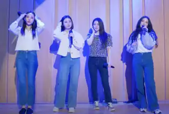 Four women standing on a stage, each holding a microphone and dressed casually in jeans and various tops, with some adjusting their hair. The stage is lit with a blue-toned light, and the background features wooden paneling.
