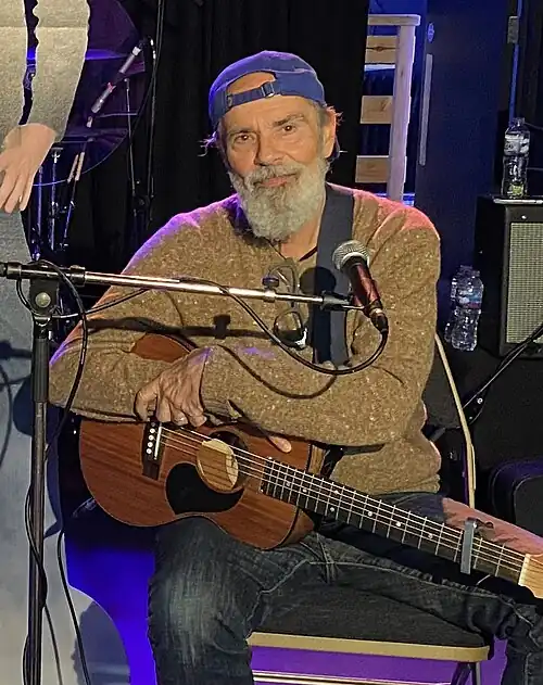 Bruce Sudano wearing a brown sweater, dark jeans, and a backwards blue cap, seated on a chair onstage in front of a microphone, holding an acoustic guitar, smiling directly at camera