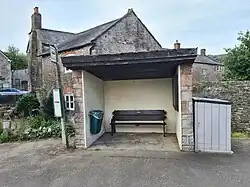 The bus shelter has a concrete floor, stone walls, and a black painted roof. There is a wooden bench inside the shelter that stands between a noticeboard on the right and a green refuse bin on the left. A bus stop sign is shown to the left of the picture and a stone house is pictured in the background.