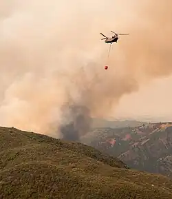 A helicopter with a bucket of water slung on a rope beneath it approaches a plume of smoke rising above scrubby green and brown hills