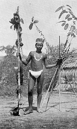 Black and white photograph of an Ocaina man in a loincloth and feathers with a pole in one hand and four long sticks in the other.