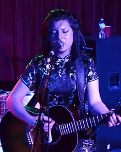 Chloe Chaidez performing on stage; singing into a microphone and playing a guitar