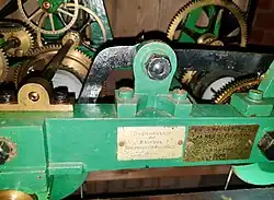 Close-up colour photograph of the clockwork installed in the tower. The metal body is painted green with a label affixed to the front that reads as follows: "Overhauled by Bristol Goldsmiths' Alliance 1939." To the right of this label is a brass plaque with the following inscription: "Repaired by James R. Cornish of Langford 1975."