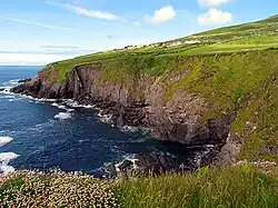 A small sea bay bordered by steep cliffs, which are topped with wild grass. On the opposite side of the bay are some green pastures surrounded by dry stone walls, and a collection of small, brightly coloured houses.