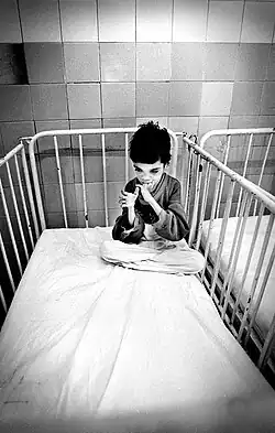 A child who appears to be very young and malnourished sits in the corner of his crib, his hands in his mouth.