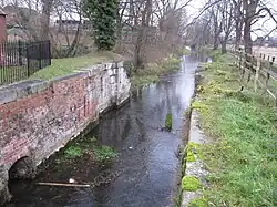 View downstream of the lock chamber build for the Sleaford Navigation, to maintain the head at the mill. The chamber is largely brick built with stone details for load-bearing parts, and the brick is coloured with moss and lichen. A little desultory grass covers the top sides. There are no lower gates, the lock having been converted into a weir many years ago. A cheap iron railing fence, painted black recently, delineates the property associated with the mill and restaurant to the left. This is a winter view and many bare trees line the banks downstream. The trunks of the nearest can be clearly seen to be covered in ivy. The water looks clear and placid.