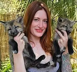 Photo of Mikayla Raines, a woman with long reddish brown hair. She holds two gray fox kits while smiling at us.