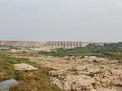 A brown dam in the distance, with a sparse field in front of it. There are wild grass, large rocks, and a small lake in the field.