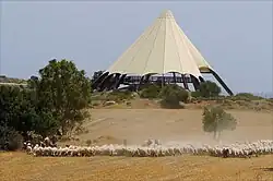 A large sand-coloured conical tent on a small hill before a pale blue sky, with a few trees and a herd of sheep on dry grassland in the foreground