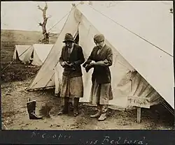 Sepia toned image of two women standing in front of a tent, both wearing below the knee skirts, high boots, jackets, and hats. They are both in the process of cleaning boots