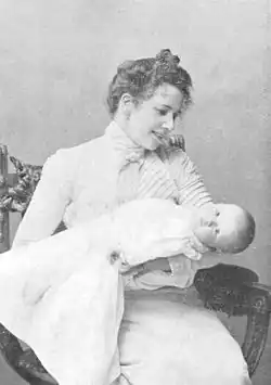 Black-and-white photo of Fohström holding a baby wrapped in a blanket. She smiles down at the baby while the baby looks at the camera.