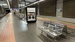 Underground grey tiled platform surrounded by concrete walls on either side. Light is coming down from above. There is a lift and set of stairs leading up to ground level.