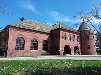 A two-story brick building with rounded windows and a tower stands on a sunny day under a mostly clear blue sky.