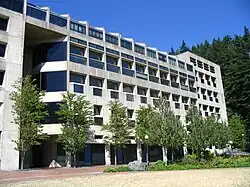 A large modern concrete university building. A plaza and strip of flora is in the foreground, while a large, tree-covered hill rises behind it.