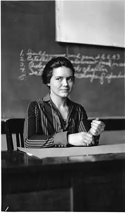 A young white woman with dark hair and eyes, seated at a table or desk indoors, wearing a striped blouse