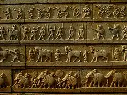 A stone depiction of four horizontal rows: the top row contains dancers, the second row contains warriors, the third contains horses and their trainers, and the bottom row depicts elephants.