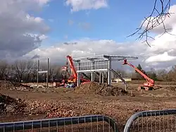 A muddy construction site with two vehicles working to build the stadium during the day. A small steel structure has been created which is beginning to resemble a football stand. The sky is lightly cloudy and in the background there are trees.