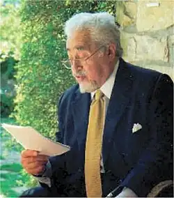 elderly white man, with neat white moustache and beard and neat full head of white hear, seated, reding a letter or other document, held in his right hand.